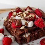 Freshly made Nutella French toast served on a white plate. It is topped with mini marshmallows, fresh raspberries, chopped hazelnuts and drizzled with extra Nutella.
