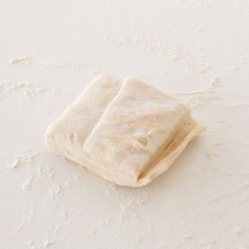 A square of puff pastry is sitting on a floured, white countertop.