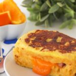 A French toast sandwich, stuffed with ham and melted cheese, sits on a white plate. A green plant and a bowl of orange segments are in the background.