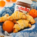 Homemade Nutella croissants are sitting in a basket lined with a blue napkin, alongside fresh apricots and a jar of Nutella.