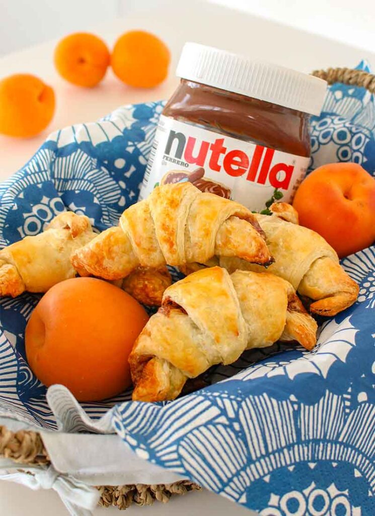 Homemade Nutella croissants are sitting in a basket lined with a blue napkin, alongside fresh apricots and a jar of Nutella.