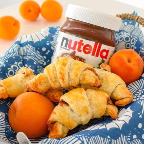 Homemade Nutella croissants are sitting in a basket lined with a blue napkin, alongside fresh apricots and a jar of Nutella.