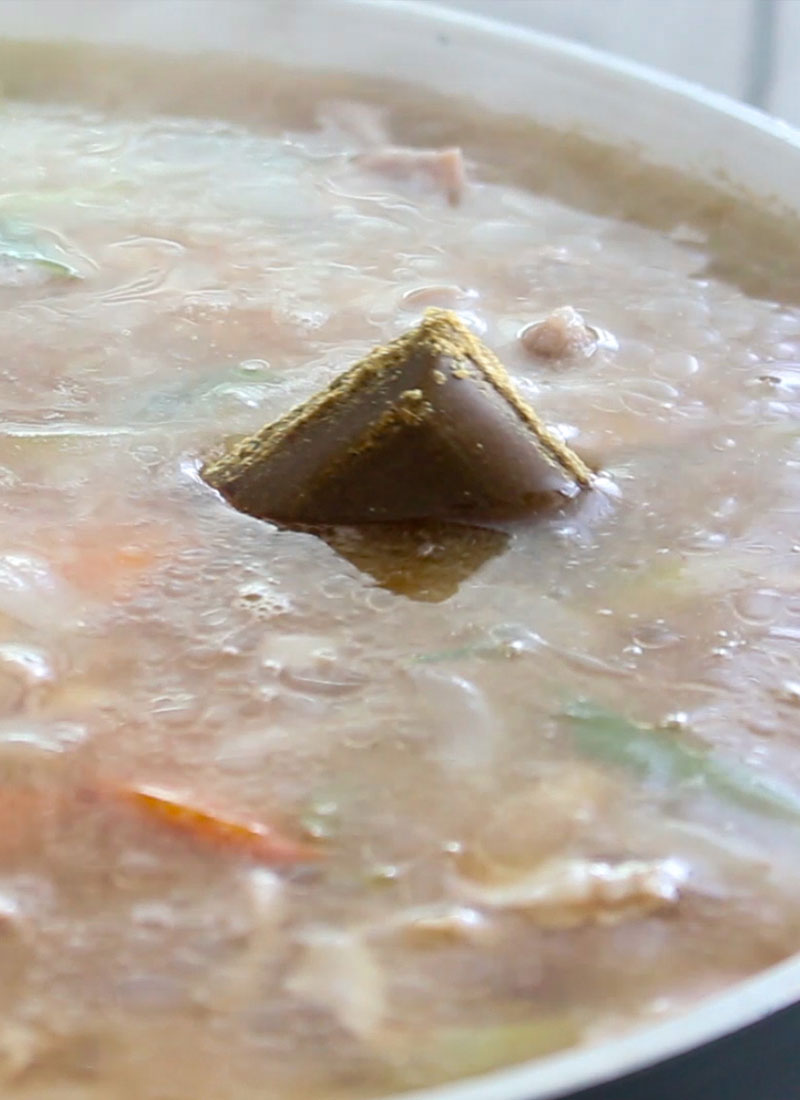 A cube of curry paste is sinking into a pot containing vegetables and stock.