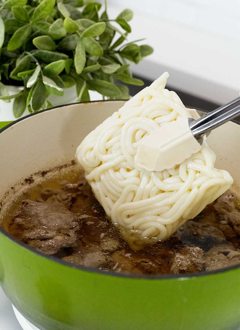 fresh udon noodles are added into a pot containing beef and soup stock