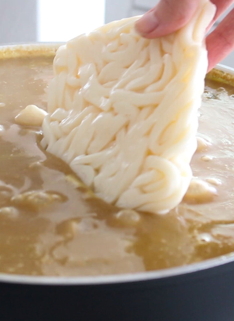 A hand is lowering some udon into a pot of bubbling curry.