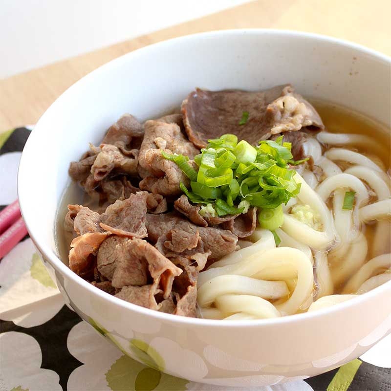 udon noodles and thinly sliced beef in a broth, topped with green onions, in a white soup bowl on a wooden table