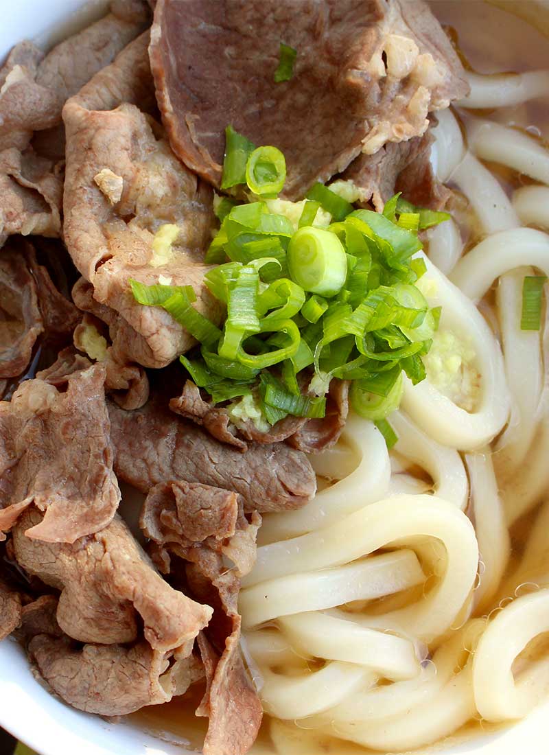 thinly sliced beef, udon noodles and green onion in a white bowl