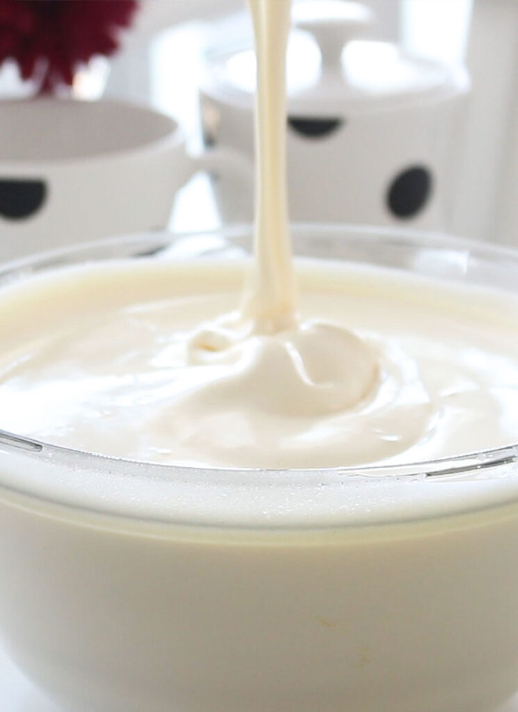 A ribbon of beaten egg and sugar mixture is falling into a large glass bowl containing more of the egg and sugar mixture.