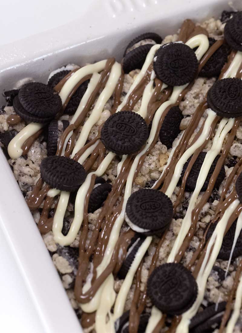 rice krispie treats in a white baking dish, drizzled with milk and white chocolate, topped with mini oreo cookies