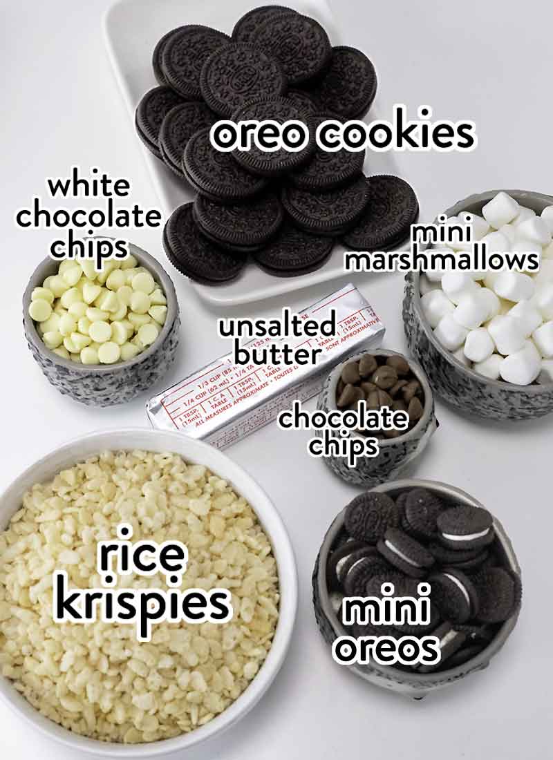 rice krispies, chocolate chips, mini marshmallows, oreo cookies and a stick of butter are in bowls and placed on a white table