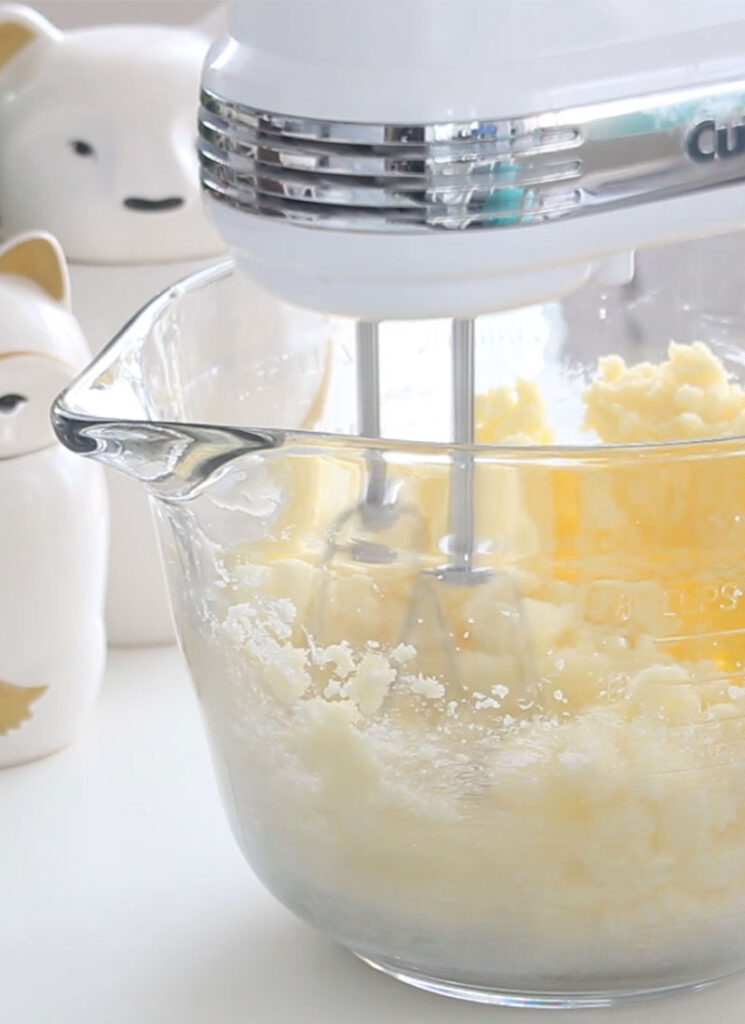 Butter and sugar is getting creamed in a large glass bowl with an electric mixer.