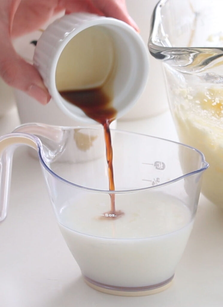 Vanilla extract is being poured into a small measuring cup containing buttermilk.