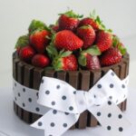 A chocolate Kit Kat cake topped with fresh strawberries and decorated with a white polka dot bow. It sits on a white surface.