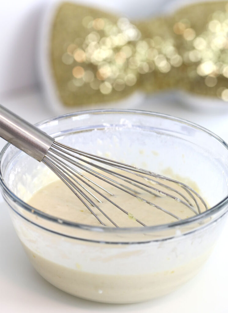 A glass bowl rests on a white countertop containing a wire whisk and pancake batter.