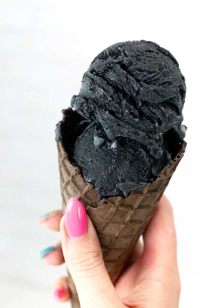 A hand holding a chocolate ice cream cone filled with two scoops of black charcoal ice cream.