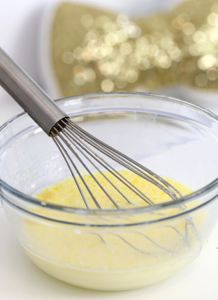 The wet ingredients for pancake batter are in a glass bowl, alongside a wire whisk.