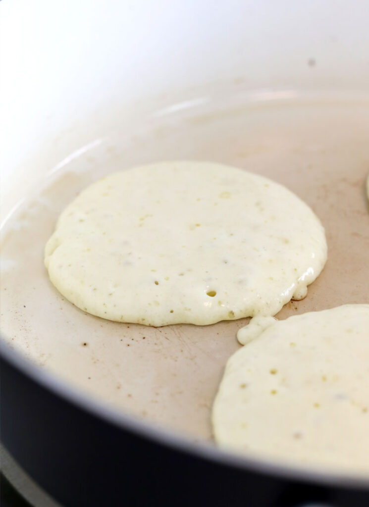 Two pancakes are cooking on a white frying pan. They have not yet been flipped.