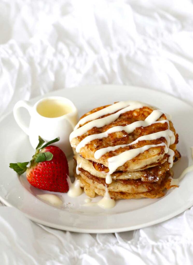A white plate containing a stack of cinnamon pancakes, a small jar of glaze and a strawberry sits on a white surface.