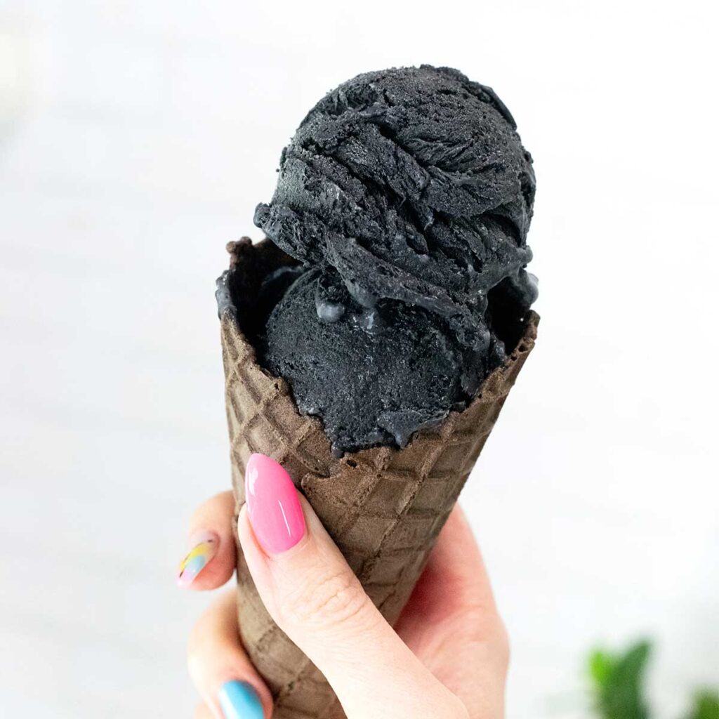 A hand holding a chocolate ice cream cone filled with two scoops of black charcoal ice cream.