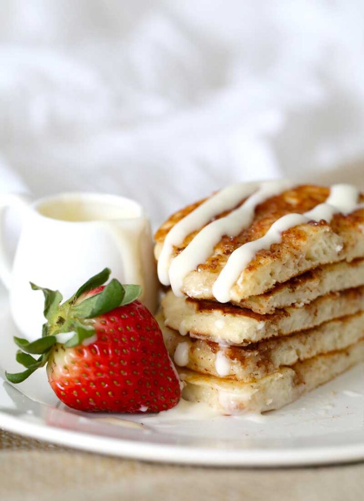 A stack of pancakes, a strawberry and a white ramekin on a white plate. The pancakes have been drizzled with cream cheese glaze.
