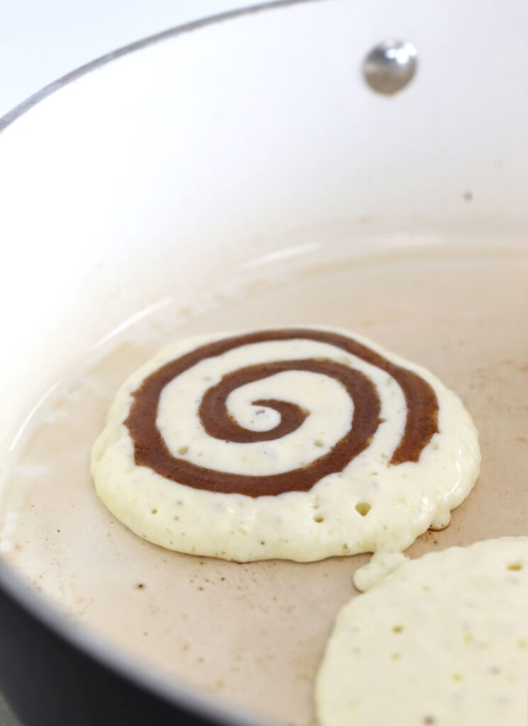 Two pancakes are cooking on a white frying pan. They have not yet been flipped. One pancake has a swirl of cinnamon piped onto the top.