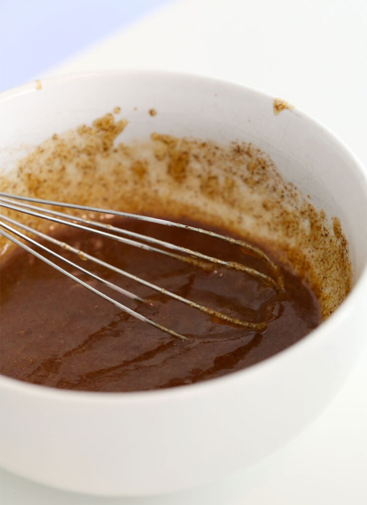 A white bowl containing cinnamon glaze and a wire whisk.