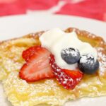 A slice of pancake is served on a white plate, garnished with whipped cream, strawberries and blueberries. It has also been dusted with powdered sugar.