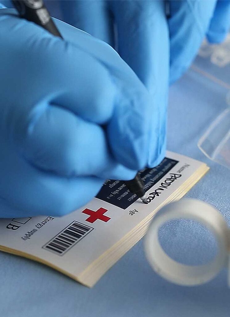 A hand wearing blue surgical gloves is writing the name on a label for a blood bag.