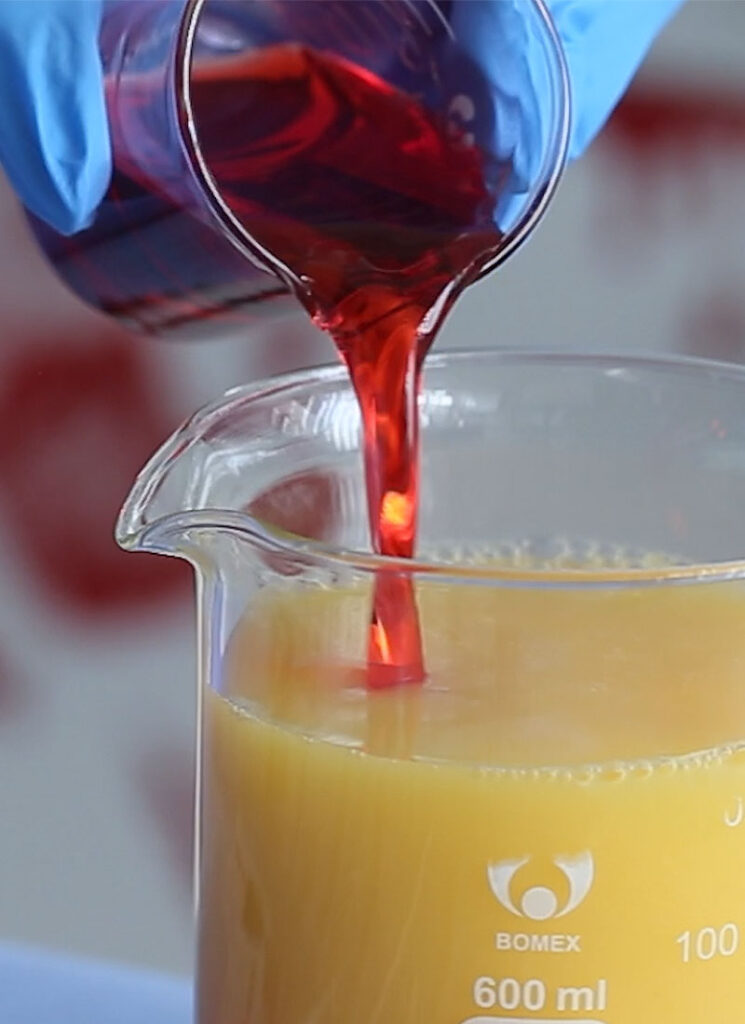 Red grenadine is being poured into a glass beaker filled with orange juice.