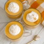 3 glasses filled with orange panna cotta and whipped cream are on a wooden tabletop, next to an orange checkered towel and a golden spoon.