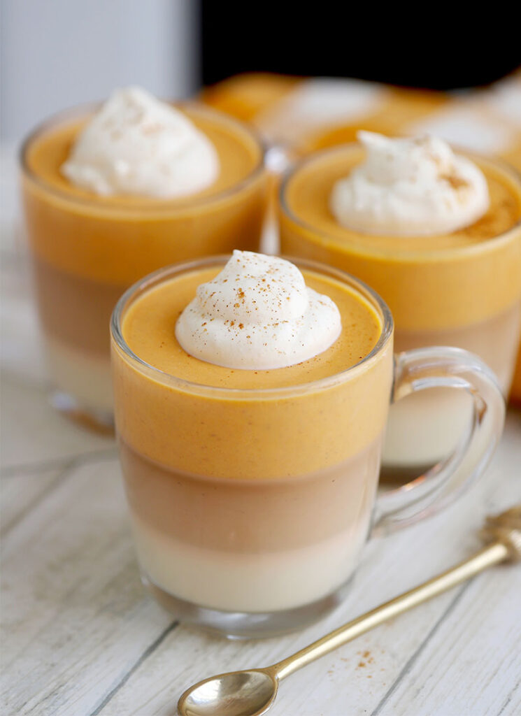 Three glasses of layered orange, brown and white panna cotta are on a wooden table with a gold spoon.