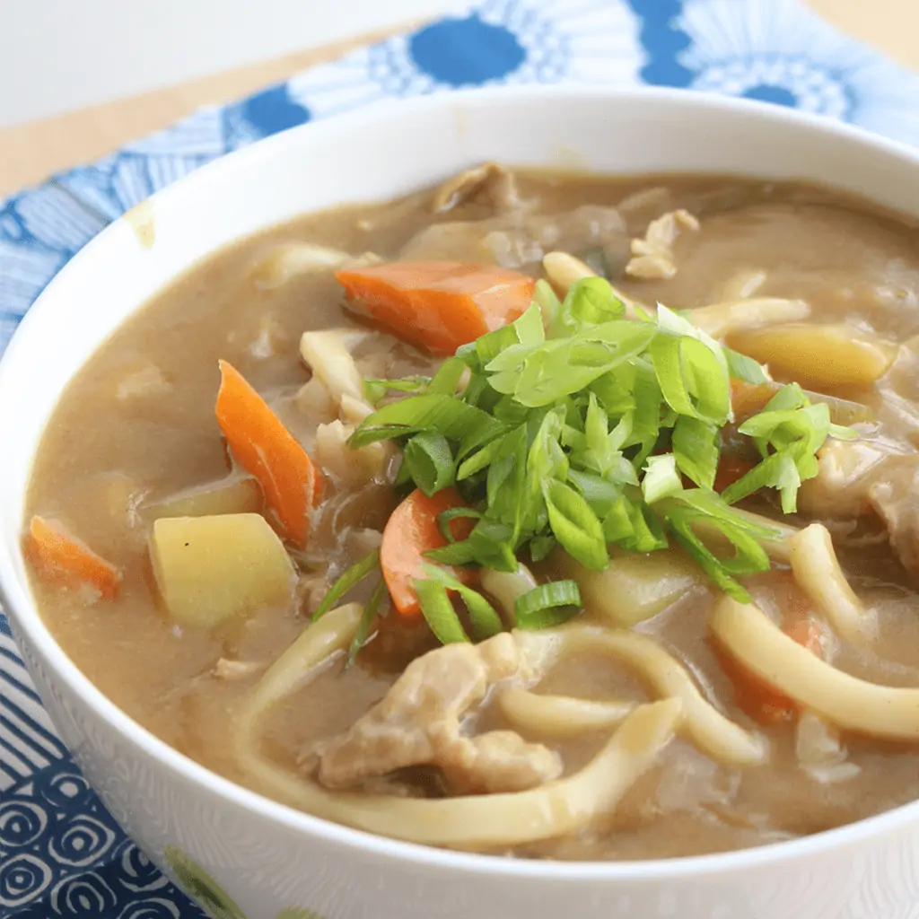 A white bowl containing curry udon, garnished with sliced green onion. The bowl is sitting on a blue and white placemat.