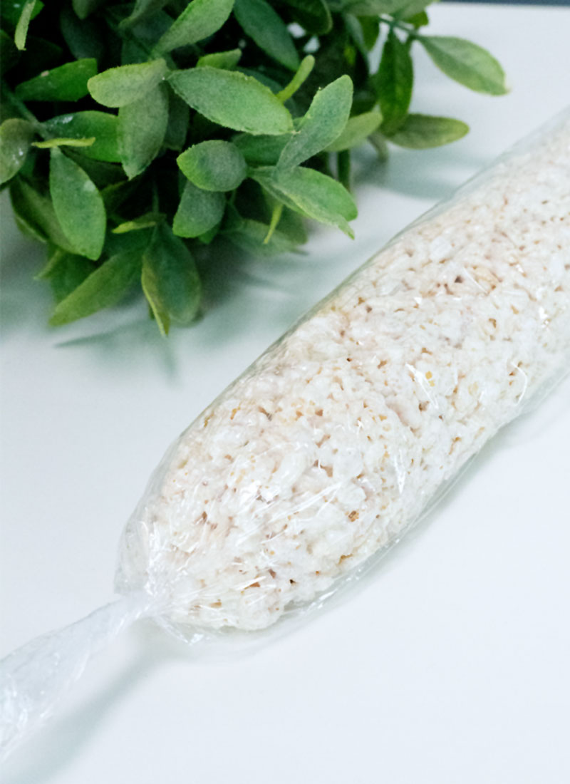 A long white rice krispie treat wrapped in plastic wrap, on a white table next to a green plant