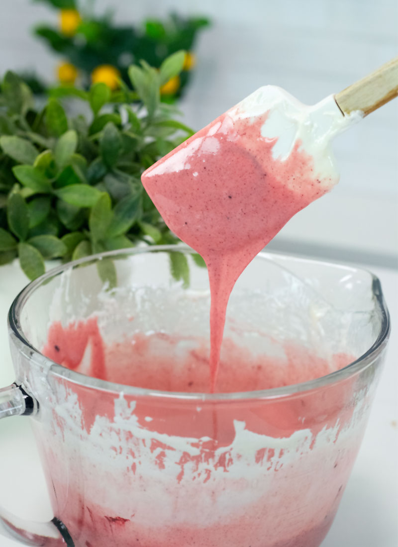 red melted marshmallow is dripping off a white spatula into a glass bowl. A green plant sits in the background