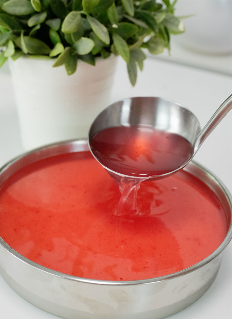 red jelly is being spooned into a round aluminum cake pan. It is on a white table with a plant