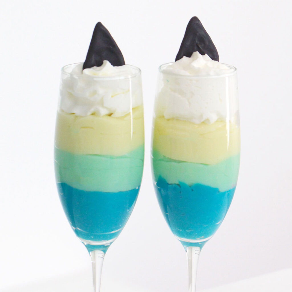 two tall glasses are filled with three layers of blue pudding, topped with a dollop of whipped cream and two black chocolate shark fins