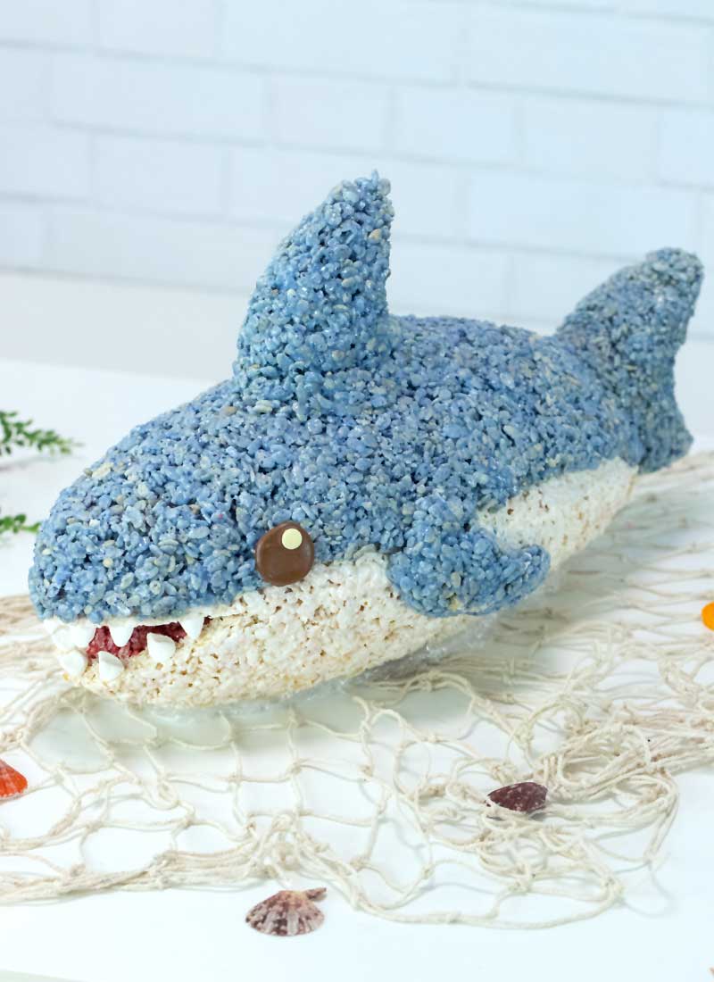 a rice krispie shark placed on a white table, on top of fishing net. Seashells and seaweed are scattered around it