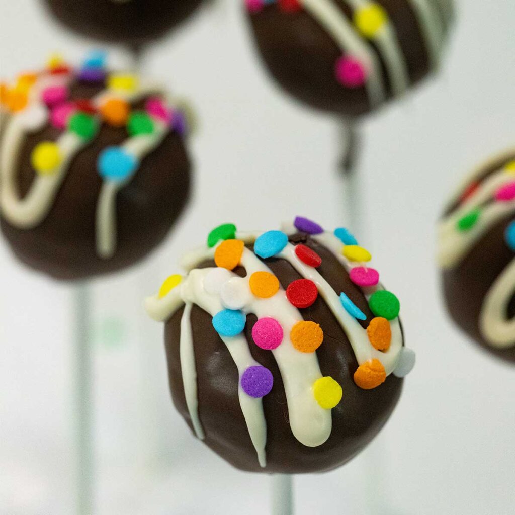 Chocolate cake pops topped with a drizzle of white chocolate and round, rainbow sprinkles are on sticks with a white background.