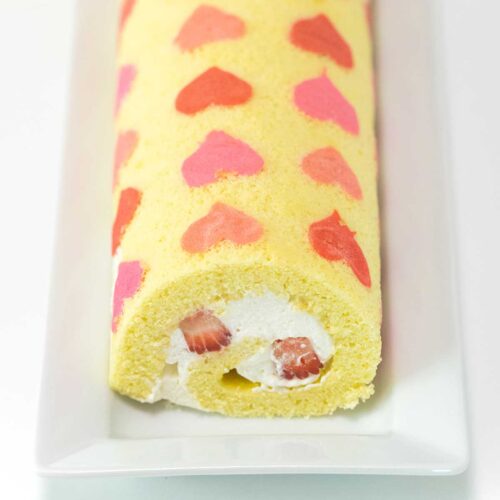 An unsliced swiss roll cake with pink and red hearts is on a long, white, rectangular dish.
