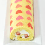 An unsliced swiss roll cake with pink and red hearts is on a long, white, rectangular dish.