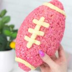A hand is holding a pink rice krispie treat football with white stitching. A white table and potted plant is in the background.