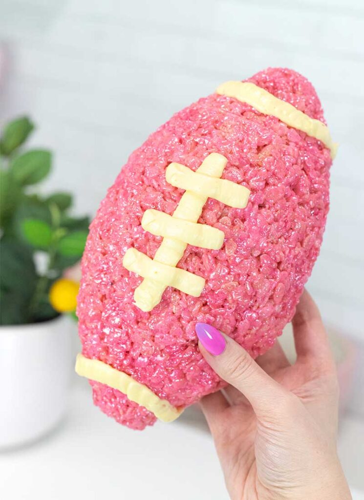 A hand is holding a pink rice krispie treat football with white stitching. A white table and potted plant is in the background.
