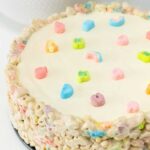 A smooth white cake has a cereal treat border. There are rainbow marshmallows in the border and on top of the cake.