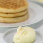 A pale yellow rabbit, made of butter, sits on a small white plate. A stack of 3 waffles sits on another plate in the background.