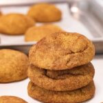 Baked snickerdoodles on a white countertop. A baking sheet containing more cookies is behind a stack of 3 cookies.