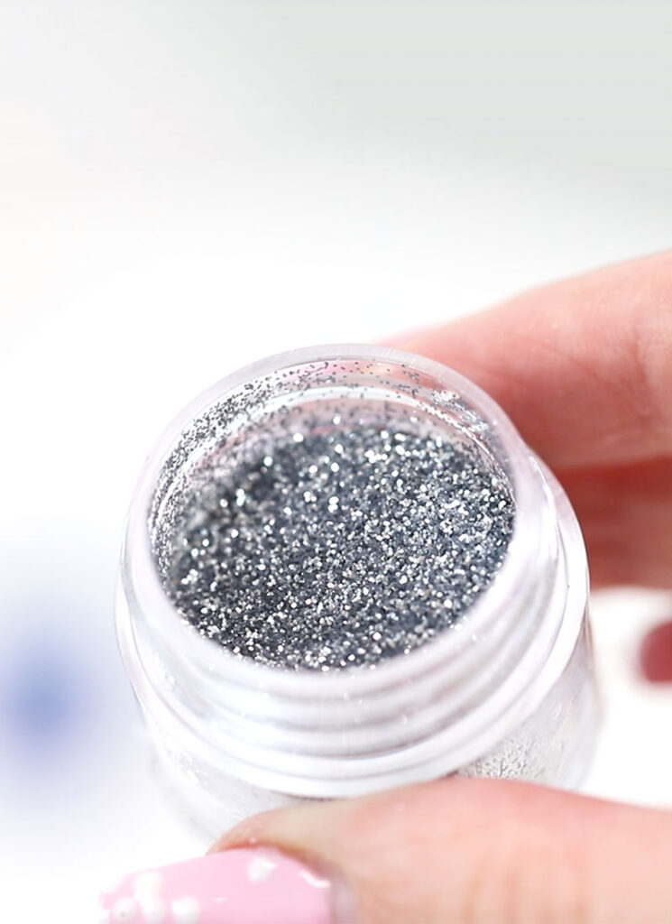 A hand is holding a small jar filled with silver glitter.