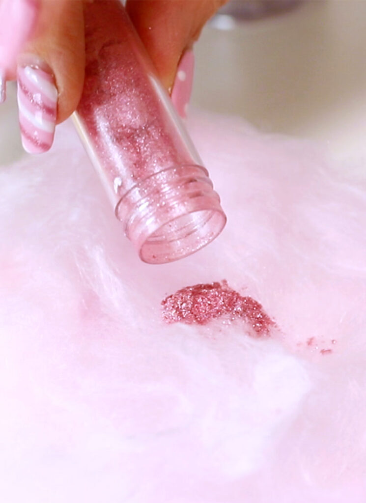 A hand is pouring pink glitter from a small clear vial onto a bed of pink cotton candy.