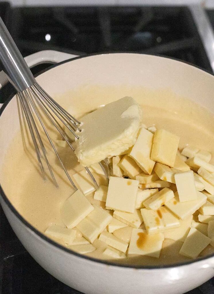 A hand is whisking a large, white pot filled with condensed milk, a large square of butter and pieces of white chocolate. The pot is sitting on a gas stove.