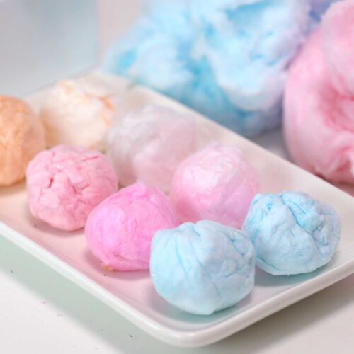A white, rectangular plate topped with 8 small balls of cotton candy, in shades of blue, pink and orange. More blue and pink cotton candy sits behind the plate, to the right.