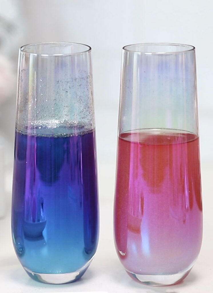 Two stemless champagne flutes are sitting on a white surface. One flute is filled with blue sparkly champagne and the other is filled with pink, sparkly champagne.